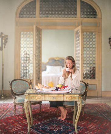 A woman relaxes with breakfast in a luxurious, classic-style hotel room with intricate details.